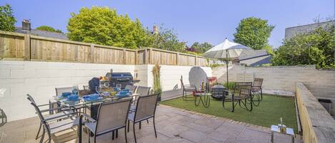 An outdoor patio with a dining table set for four, surrounded by chairs. There is a barbecue grill and a covered seating area.