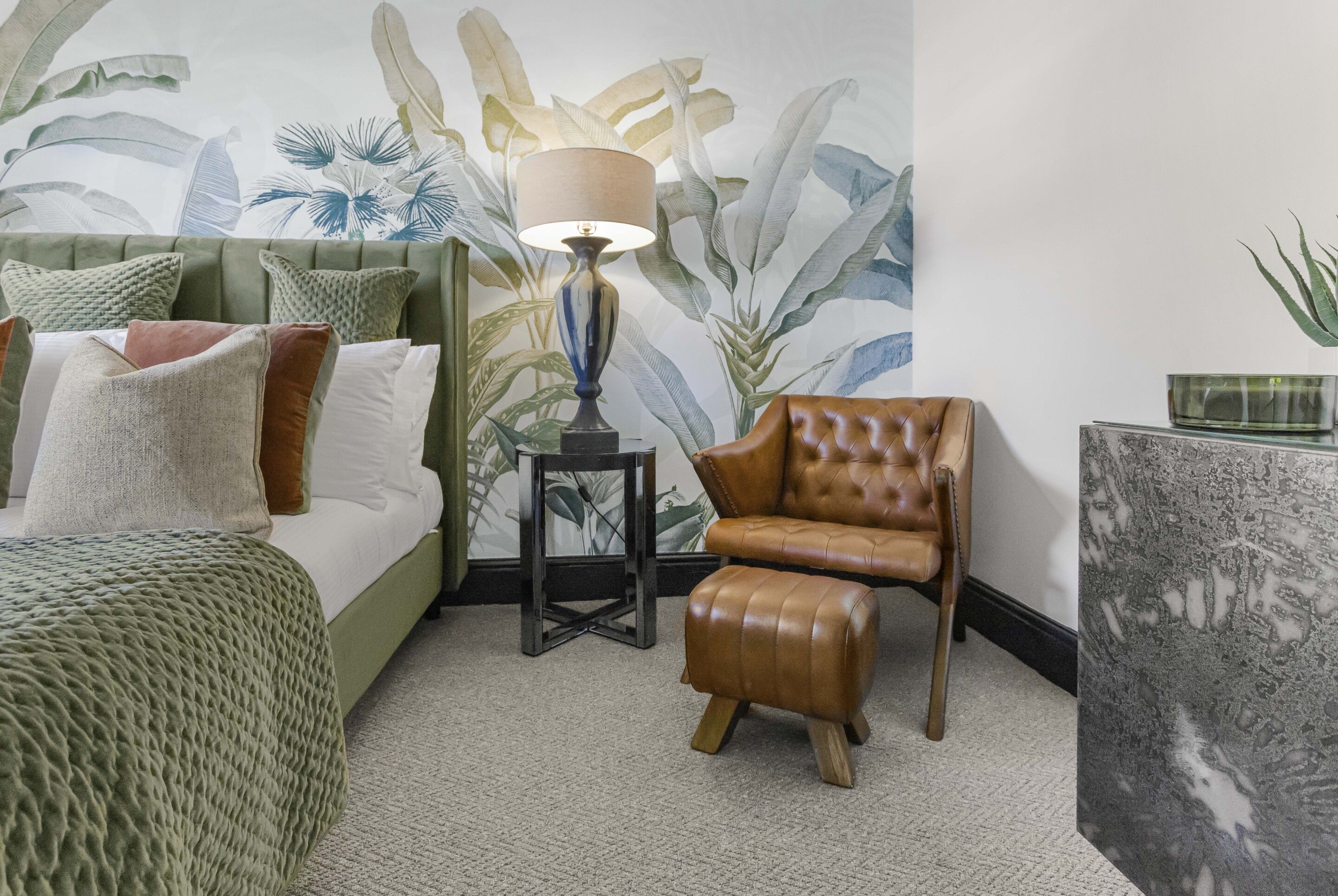 A room with a bed, a green upholstered headboard, a brown leather armchair, a small wooden stool, a floor lamp, and a wall with a tropical leaf mural.