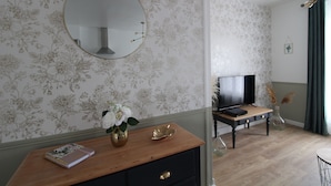 A room with floral wallpaper, a round mirror, a wooden desk with a notebook, a vase with flowers, a television, and a lamp. - Les Nuits Bonneuilloises Le Henson (Breteuil)