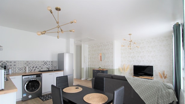 A modern kitchen with a washing machine, dining table with chairs, and a sofa with a TV. - Les Nuits Bonneuilloises Le Henson (Breteuil)