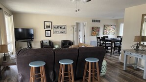 Ein Wohnzimmer mit Ecksofa, zwei Barhocker, einem Fernseher und einem Essbereich mit Tisch und Stühlen.