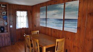 Ett matrum med träpanel, ett bord och stolar, ett fönster med spetsgardiner och en utsikt över havet som syns genom en tavla.