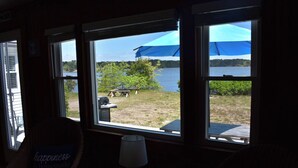Ett rum med stora fönster med utsikt över en brygga och en sjö, en flätstol med en kudde och ett bord med en lampa.