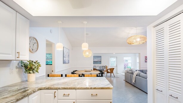A modern kitchen with white cabinetry, a marble countertop, and pendant lighting. There is a dining area with a round table and chairs, and a living room with a sofa and a TV. - Coastal Getaway minutes from Siesta Key, America’s #1 Beach (Sarasota)
