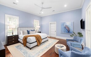 Une chambre à coucher comprenant un lit, des tables de chevet, un ventilateur de plafond, une grande fenêtre avec des stores, un téléviseur fixé au mur et un tableau.