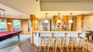 A spacious kitchen with wooden cabinets, a central island with bar stools, and a pool table in a room with large windows. - Lake Access, Dock, Hot Tub, Fire Pit , Free Golf + Pickle Ball (Swanton)