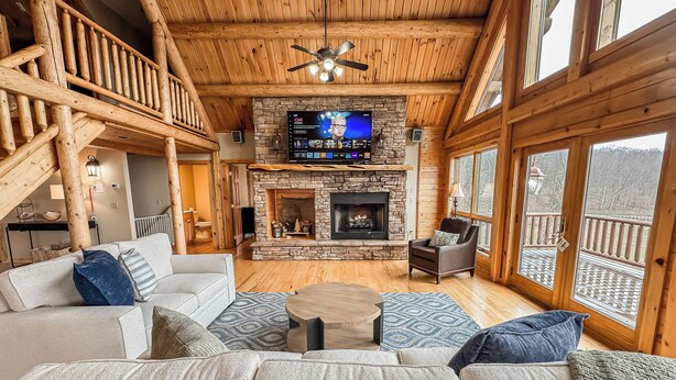 A cozy log cabin interior with a stone fireplace, wooden ceiling, and a large TV screen. - Lake Access, Dock, Hot Tub, Fire Pit , Free Golf + Pickle Ball (Swanton)