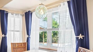 Una habitación con una mesa de comedor, dos sillas, una lámpara colgante y ventanas grandes con cortinas.
