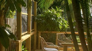 Una terraza de madera con dos sillas de mimbre y una mesita pequeña, rodeada de abundante follaje y una valla de madera.