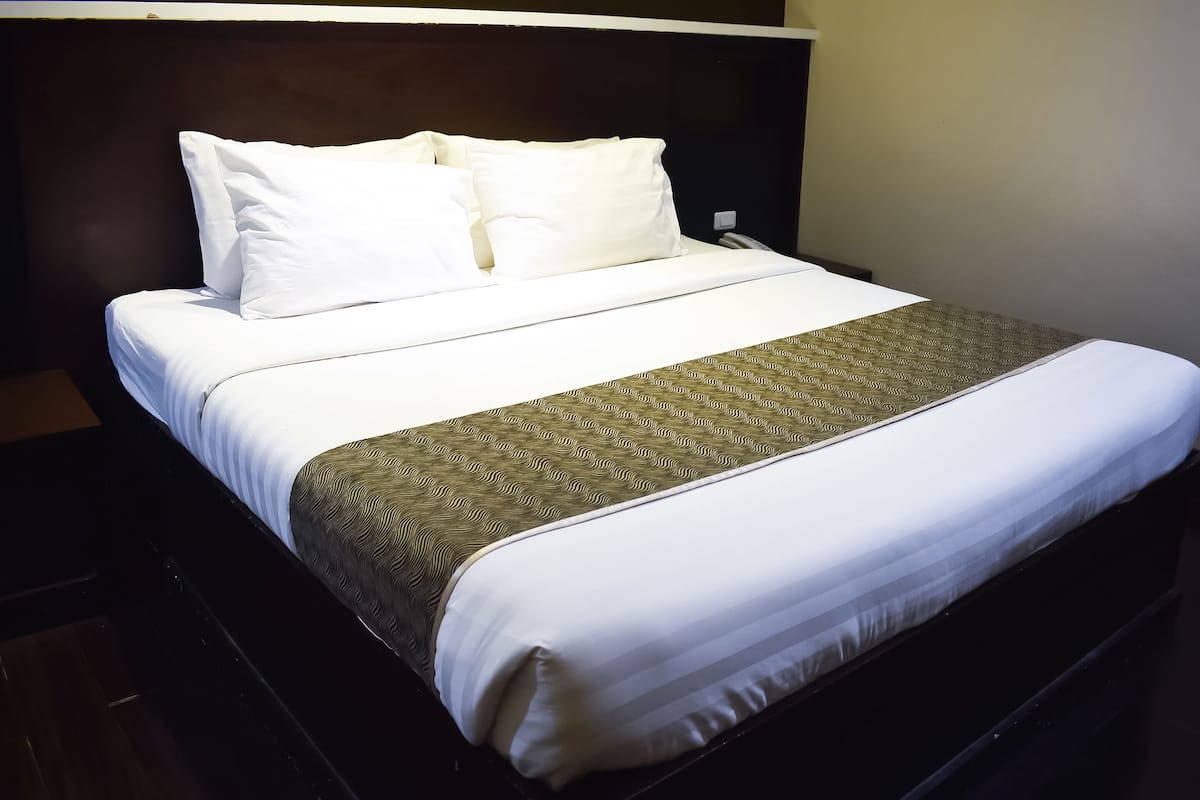 A neatly made bed with white linens and a patterned bedspread, a dark headboard, and a plain wall.