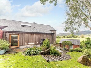 Une grange rustique en bois, dotée d’un toit en métal, d’un petit espace extérieur aménagé pour s’asseoir et d’un jardin avec un mur en pierre et un sentier.