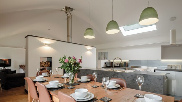 A modern dining area with a wooden table set for six, surrounded by chairs. A vase with flowers is placed on the table. - This striking home is ideally located between the beach and the hills. (Arthog)