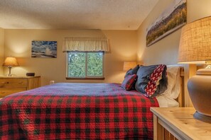 A bedroom with a bed, a window with a view, a lamp, and a painting on the wall. - The Trailhead Townhouse (West Yellowstone)