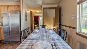 A dining area with a checkered tablecloth, a stainless steel refrigerator, and a window with blinds. - The Trailhead Townhouse (West Yellowstone)