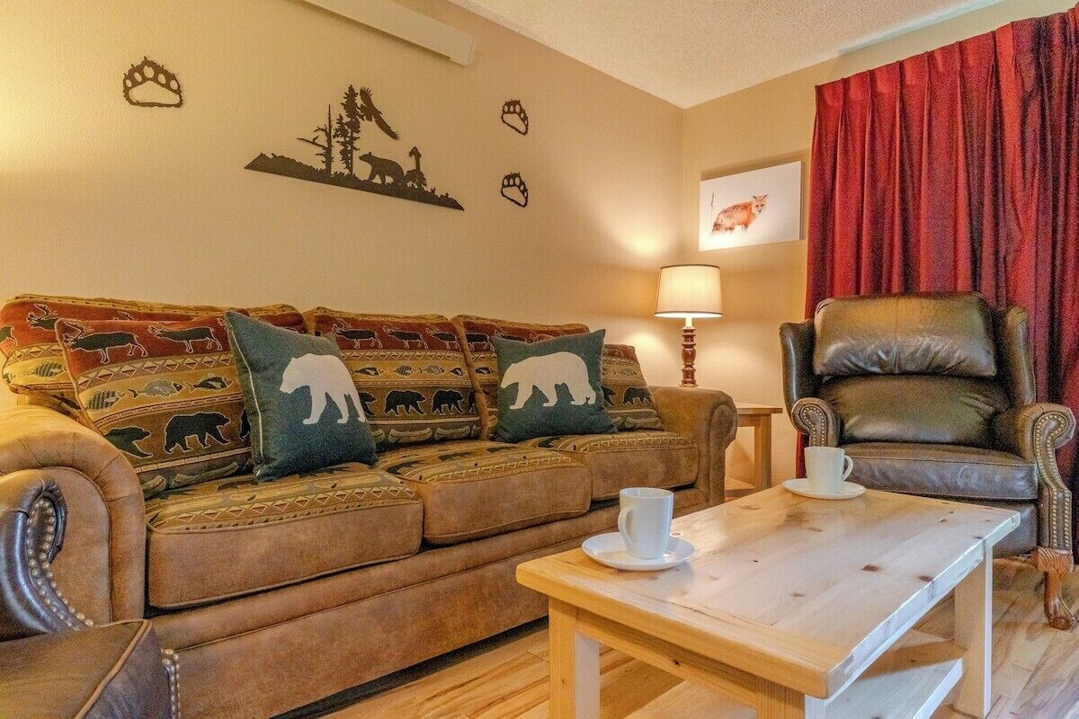 Een gezellige woonkamer met een bruine bank versierd met dierenkussens, een houten salontafel, een leren fauteuil en een rood gordijn.