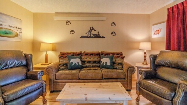 A living room with a brown sofa set, two armchairs, a coffee table, and wall art depicting animals. - The Trailhead Townhouse (West Yellowstone)