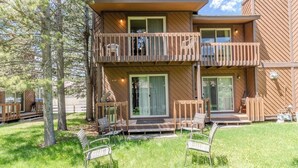 A two-story wooden cabin with a balcony, outdoor seating, and a grassy yard. - The Trailhead Townhouse (West Yellowstone)