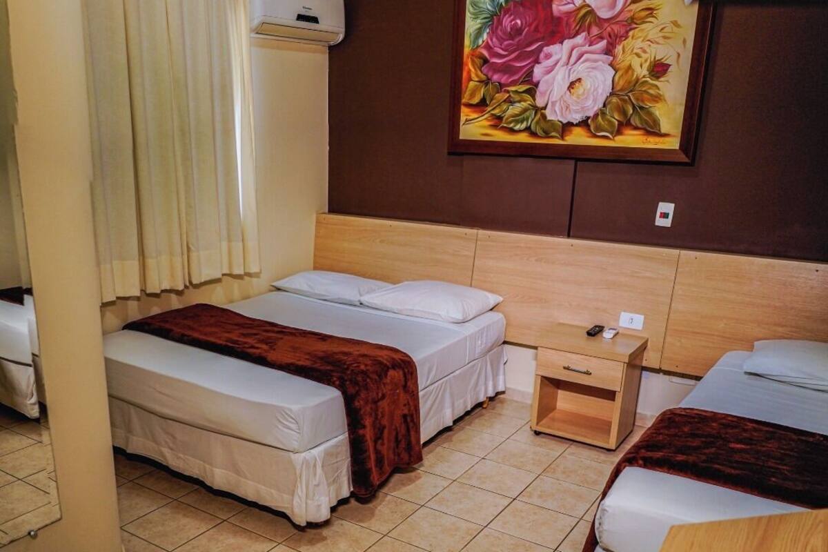 Une chambre d’hôtel avec deux lits, une tête de lit en bois, un tableau représentant des fleurs, une petite table de chevet en bois et une fenêtre avec des rideaux.