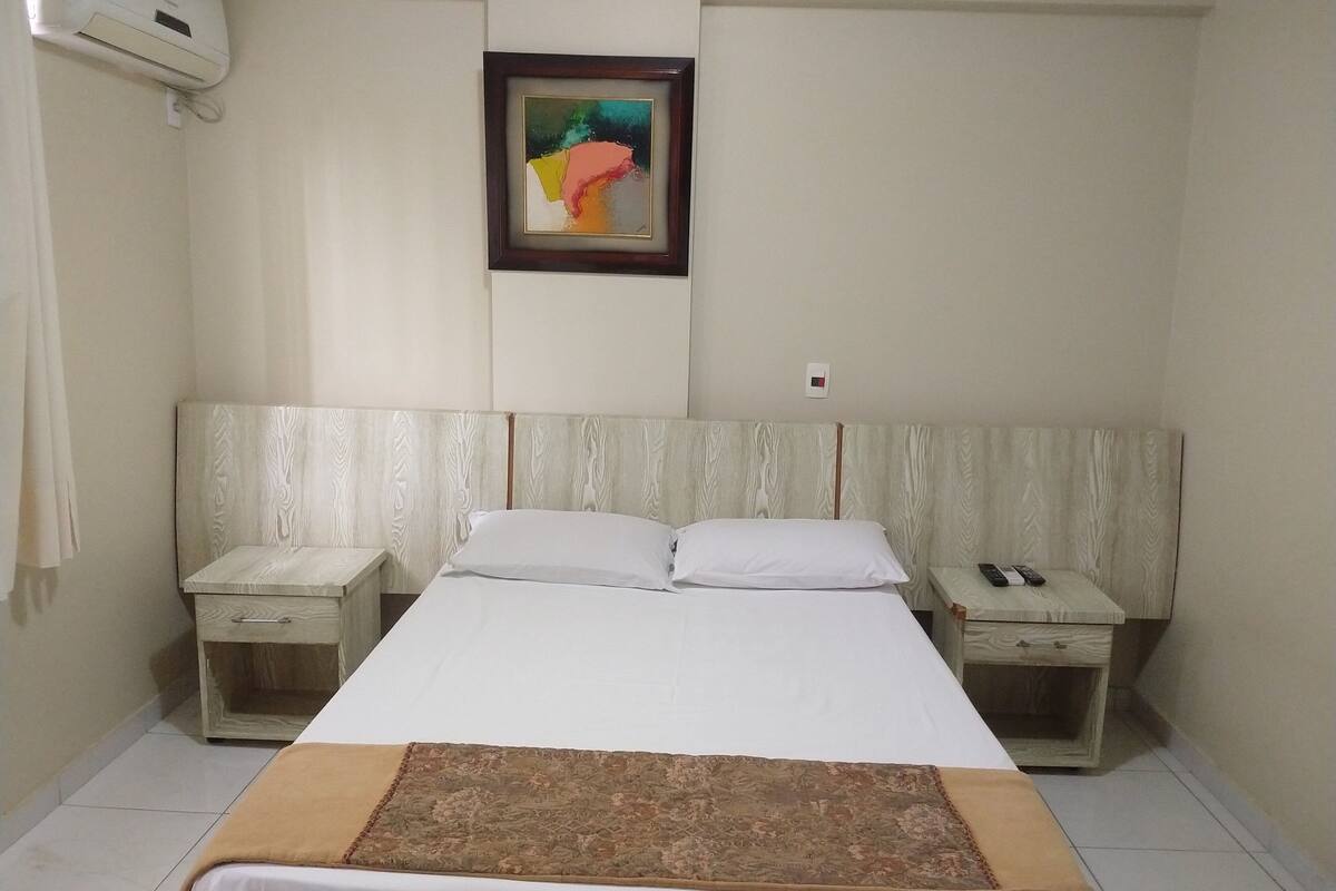 Une chambre d’hôtel comprenant un lit, deux tables de chevet, une petite table, un climatiseur fixé au mur et un tableau encadré.