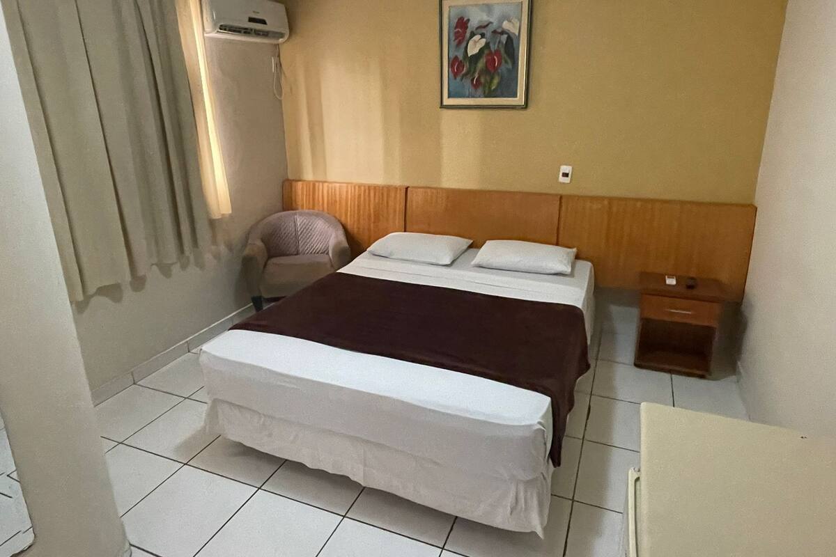 Une chambre d’hôtel avec un lit, une chaise, un appareil de climatisation, un tableau au mur et une table de chevet.