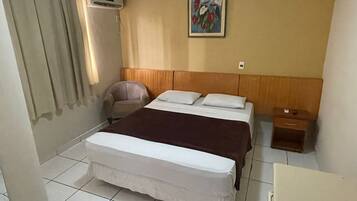 A hotel room with a bed, a chair, an air conditioning unit, a painting on the wall, and a nightstand.