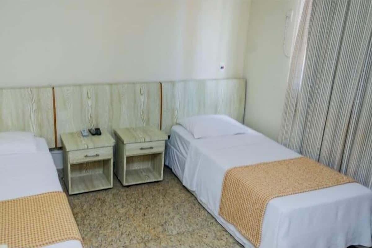 Une chambre d’hôtel avec deux lits simples, une tête de lit en bois, une petite table de chevet avec un téléphone et une fenêtre avec des stores.
