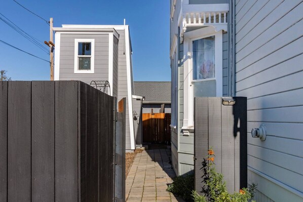 Un étroit sentier mène à une propriété résidentielle, bordée d’une clôture en bois d’un côté et d’un mur extérieur blanc de l’autre.