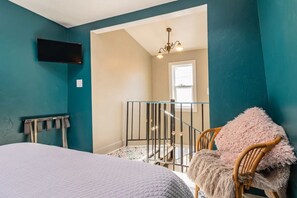 Une chambre avec des murs de couleur turquoise, un lit, une chaise recouverte d’une couverture moelleuse, un téléviseur fixé au mur et un escalier menant à une autre pièce.