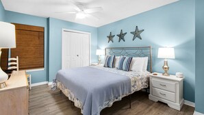 A bedroom with a bed, nightstands, a desk, a chair, a ceiling fan, and wall decorations. - Cute 2/2 condo - 2 KING BEDS, Queen Sofa Sleeper and hall bunks (Destin)