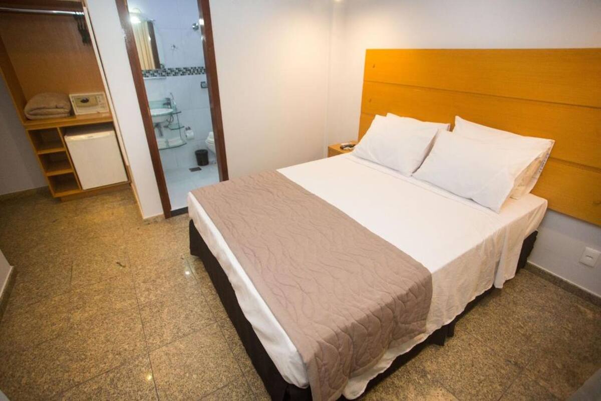 Una cama bien hecha con cabecera de madera, una mesita de noche y un baño visible a través de una puerta abierta.