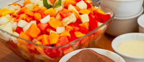Eine Schüssel Obstsalat mit gewürfelten Früchten und Minzblättern, umgeben von Schüsseln mit verschiedenen Zutaten wie Kakaopulver und einer cremigen Masse.