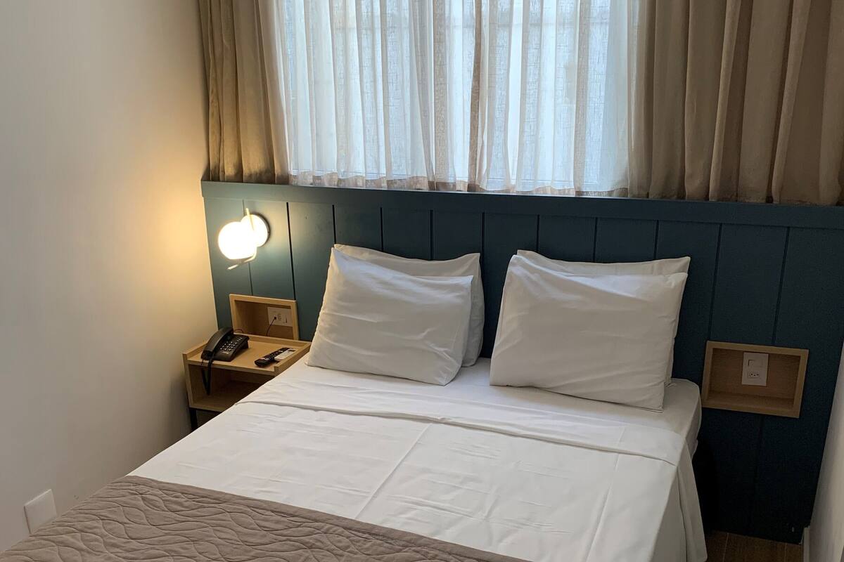 Una cama bien hecha con una colcha acolchada, dos almohadas, una lámpara de noche y una mesita de noche con un teléfono.