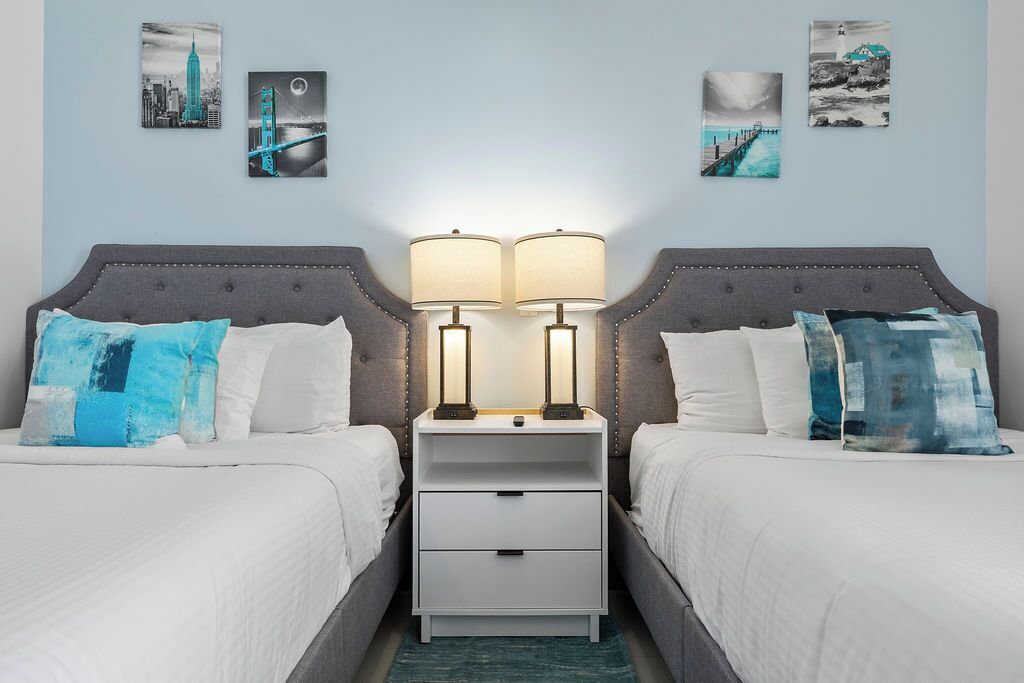 A hotel room with two beds, each with a grey headboard and white bedding. There are two bedside tables with lamps, a white nightstand with a drawer, and three wall-mounted pictures depicting cityscapes and a lighthouse.