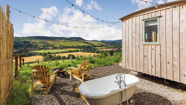 A wooden cabin with a bathtub and two Adirondack chairs overlooking a scenic landscape. - Woodside Hut 3 - UK36107 (Strathpeffer)
