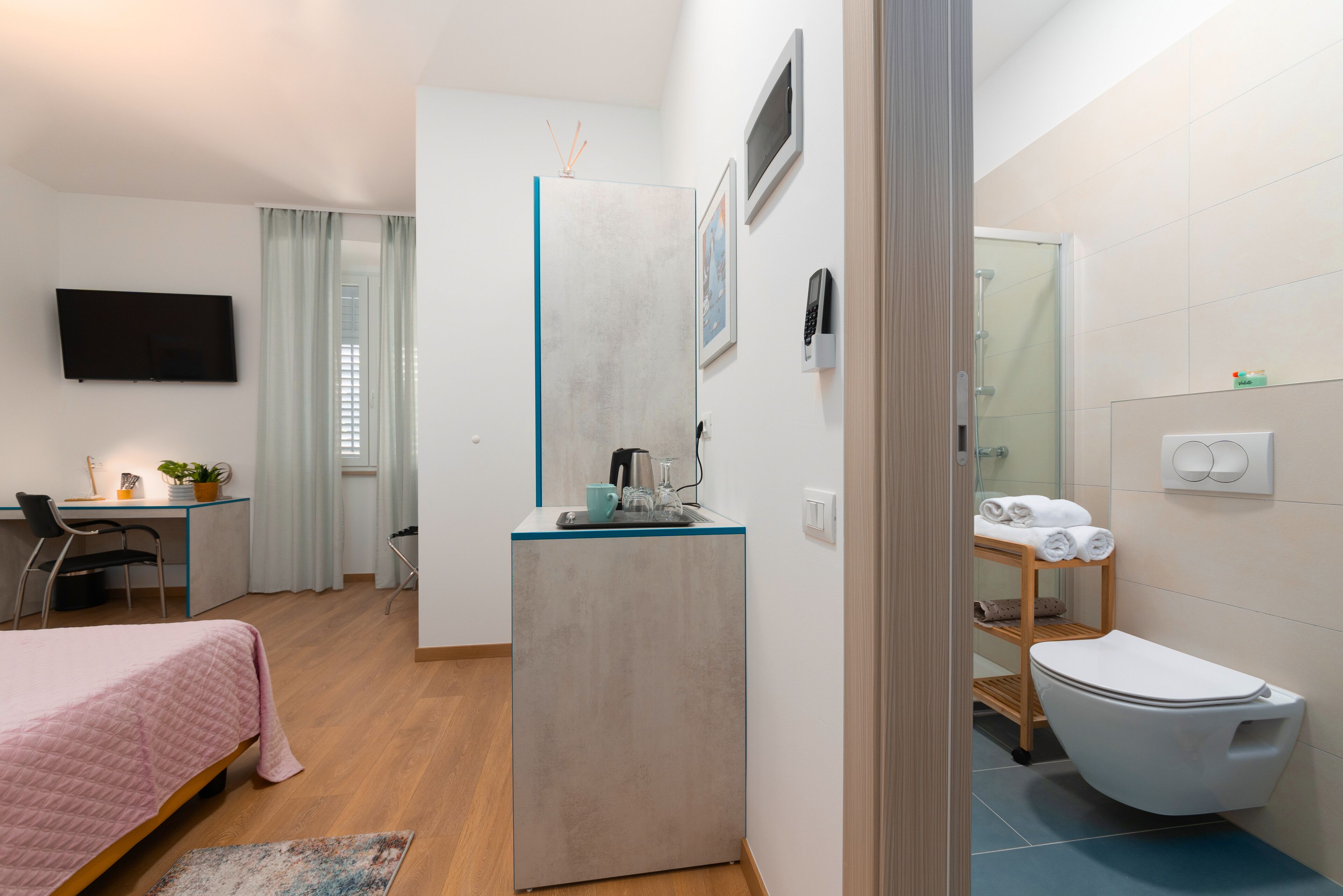 Habitación de hotel pequeña con cama, escritorio, kitchenette, baño con inodoro y ducha, y televisor montado en la pared.
