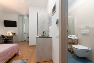 Quarto de hotel compacto com cama, escrivaninha, pequena cozinha e banheiro com vaso sanitário e chuveiro, além de televisão presa na parede.
