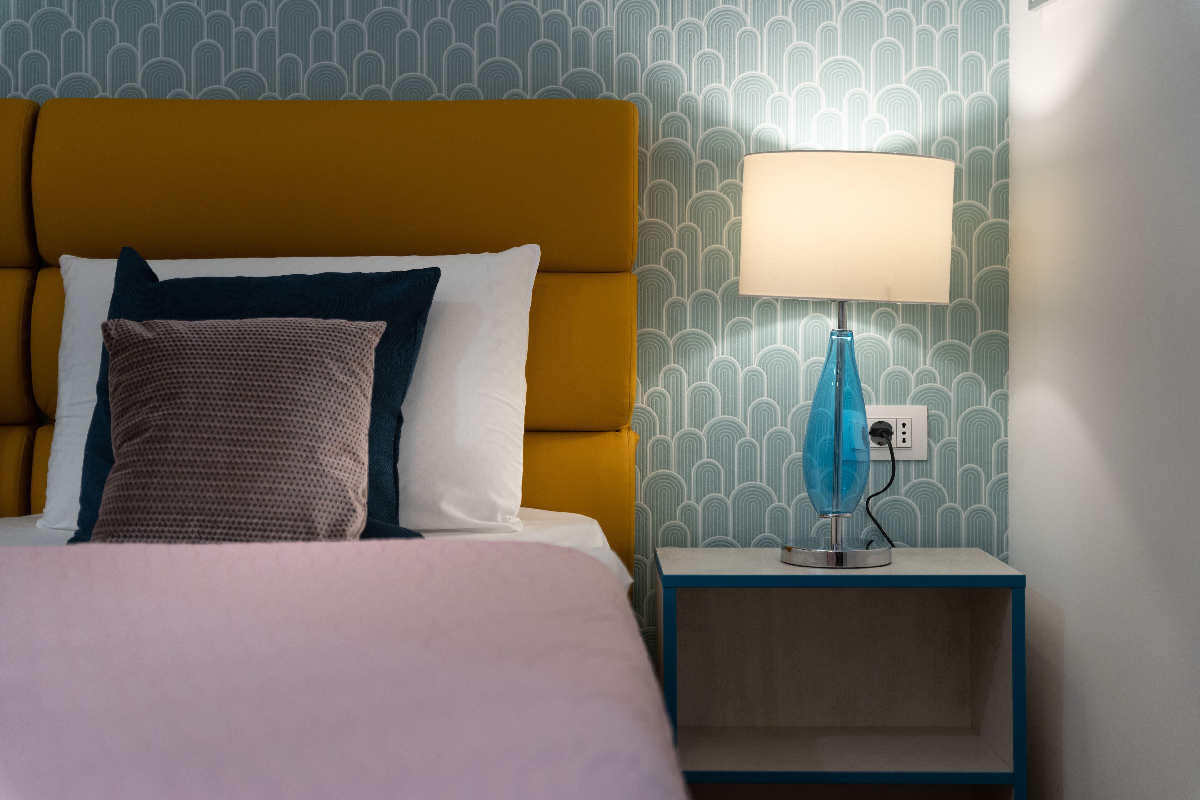 Een net opgemaakt bed met een mosterdkleurig hoofdeinde, een blauwe bedlamp en behang met een patroon.