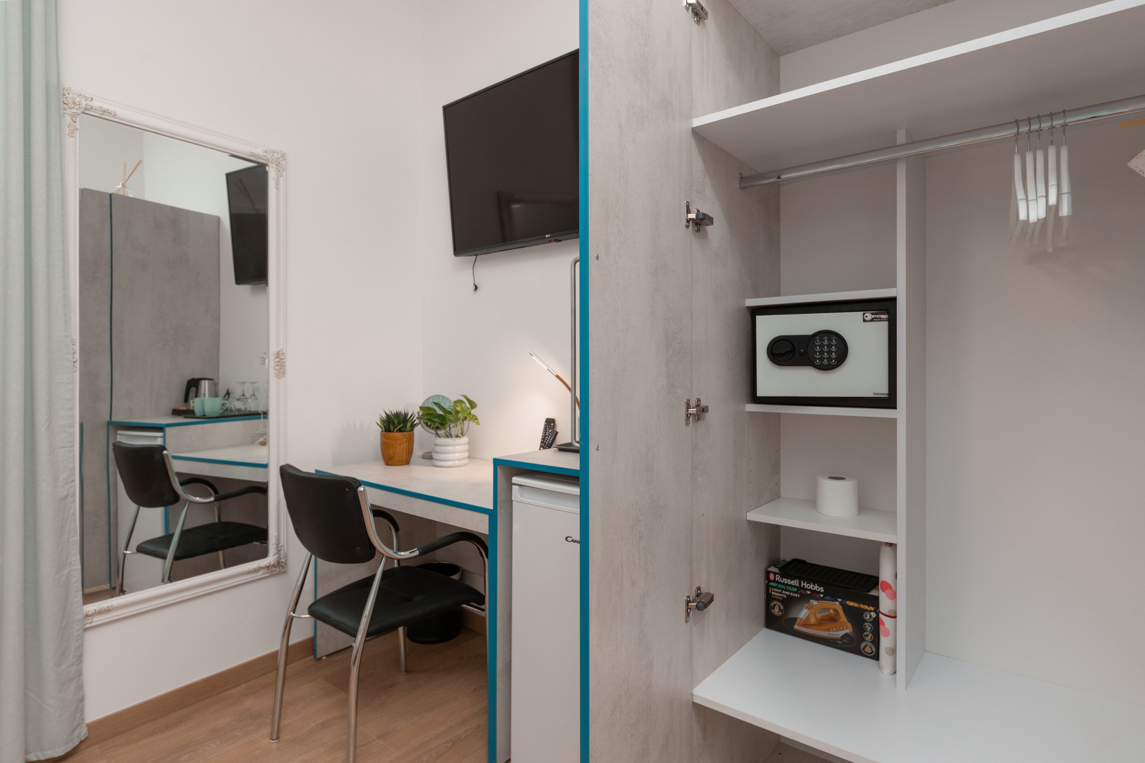 A compact room with a desk, chair, and a small refrigerator. A television is mounted on the wall, and there is a shelf with a microwave and a box of tissues.