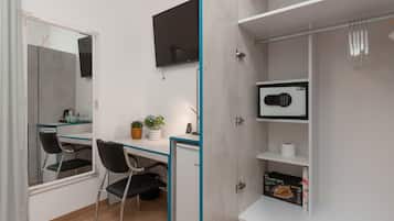 A compact room with a desk, chair, and a small refrigerator. A television is mounted on the wall, and there is a shelf with a microwave and a box of tissues.