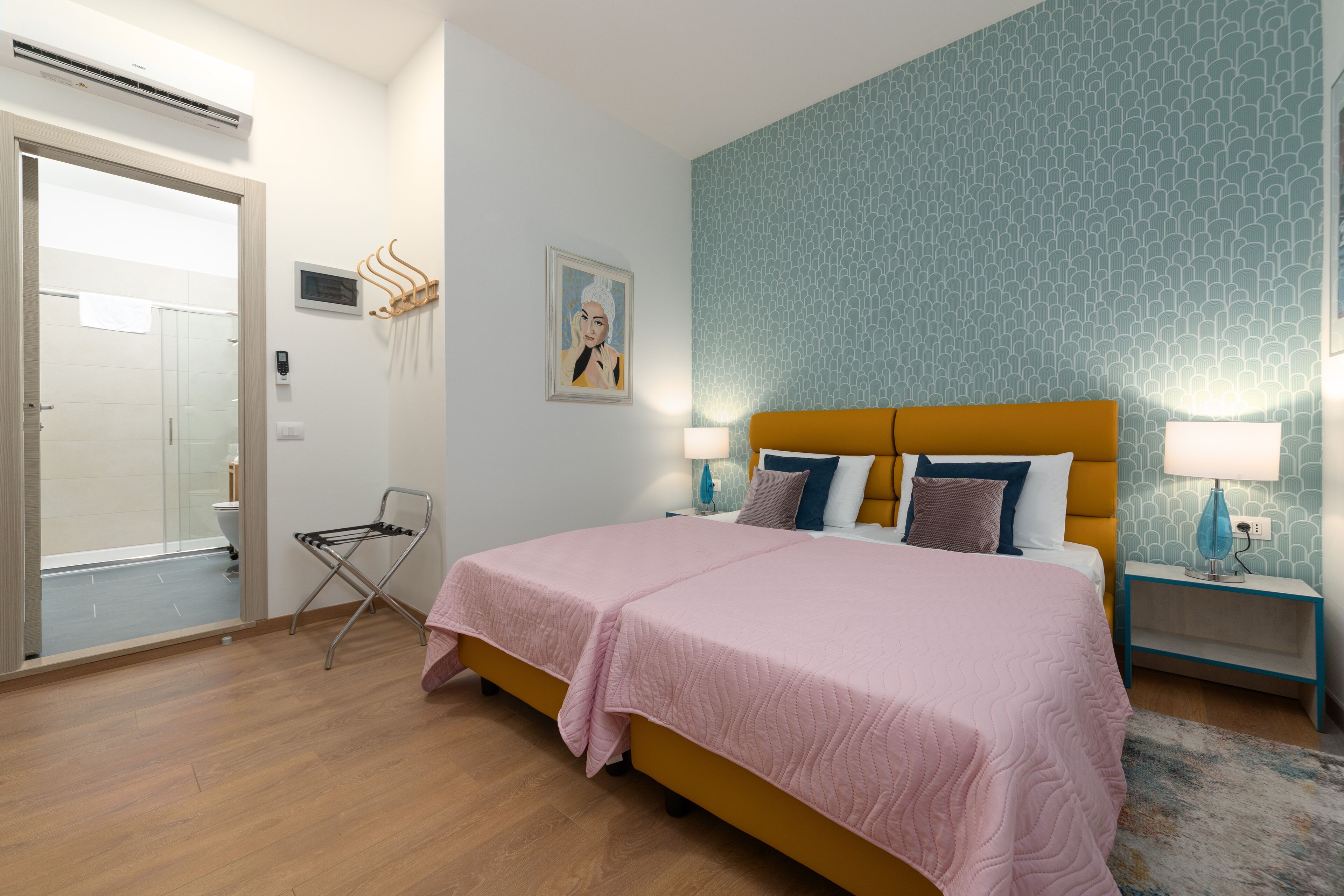 A bedroom with a double bed, a nightstand with a lamp, a wall-mounted towel rack, and a door leading to a bathroom.
