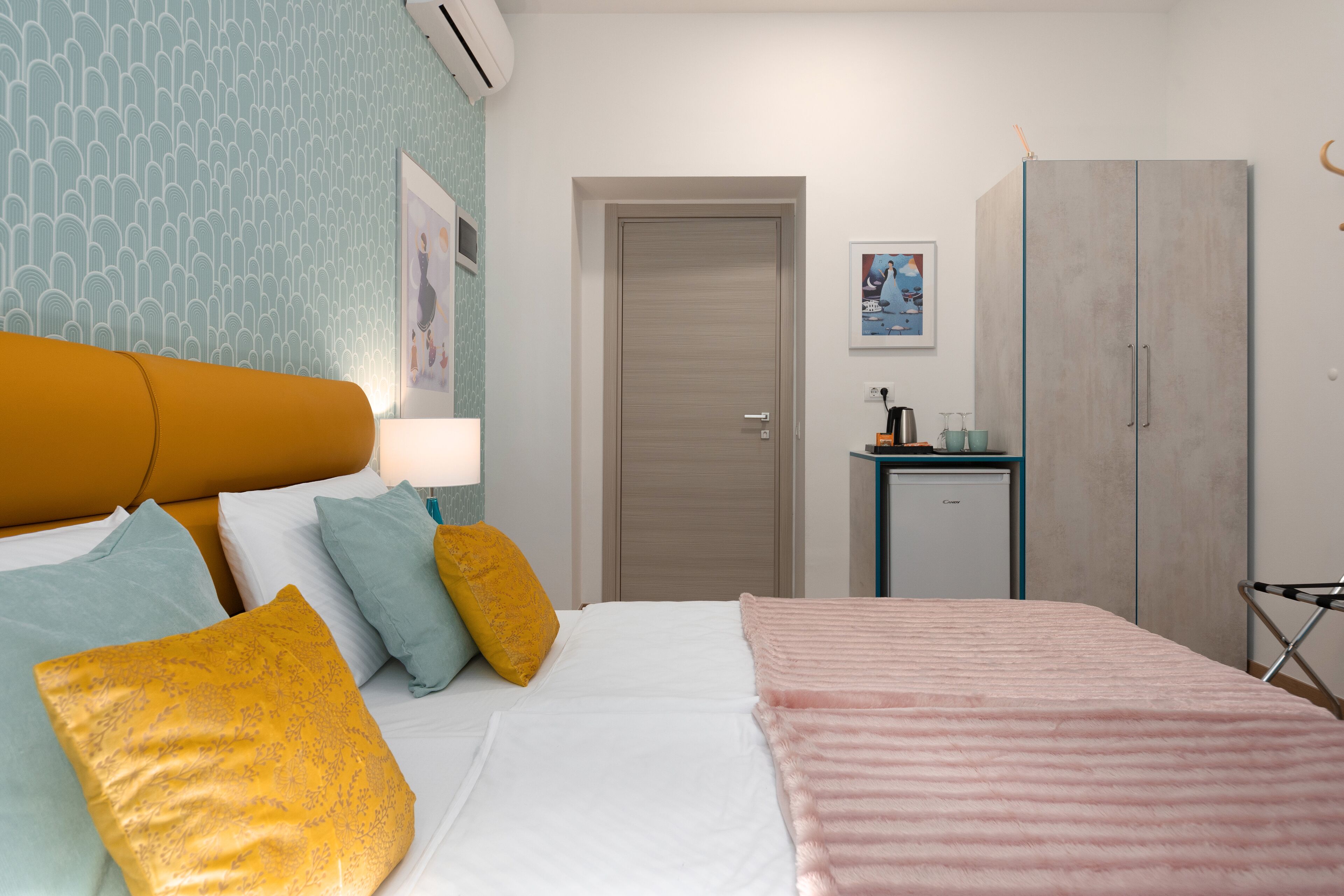 A hotel room with a bed, pillows, a bedside table with a lamp, a wardrobe, and a wall with a patterned wallpaper.