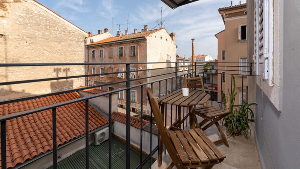 A balcony with a wooden table and chairs, a potted plant, and a view of neighboring buildings. - Polesana Rooms (Pula)