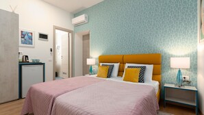 A hotel room with a double bed, two bedside tables, a small desk with a lamp, a refrigerator, and a wall-mounted air conditioner. - Polesana Rooms (Pula)