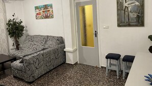 Una stanza con un divano a fantasia, un tavolino, due sgabelli, una pianta in vaso, un quadro e un condizionatore d'aria.