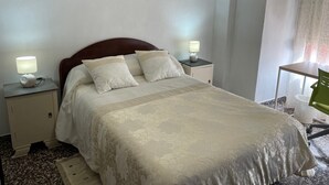 Un letto rifatto con cura, una testiera in legno, due lampade sui comodini e un tavolino con una sedia. Sopra il letto, tre opere d'arte incorniciate.