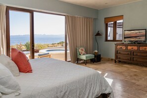 A bedroom with a large bed, a chair, a dresser, and a TV. There is a view of the sea through the window.