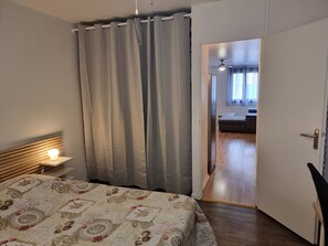 Makuuhuone, jossa on sänky, yöpöytälamppu, verho ja ovi, joka johtaa toiseen huoneeseen.