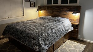 Quarto com cabeceira de madeira, colcha estampada com flores e janela com persianas.