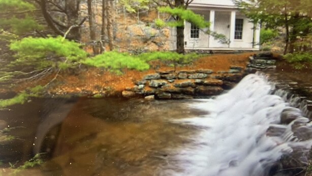 Uma casa com uma ponte de pedra sobre um riacho, cercada por árvores e vegetação.