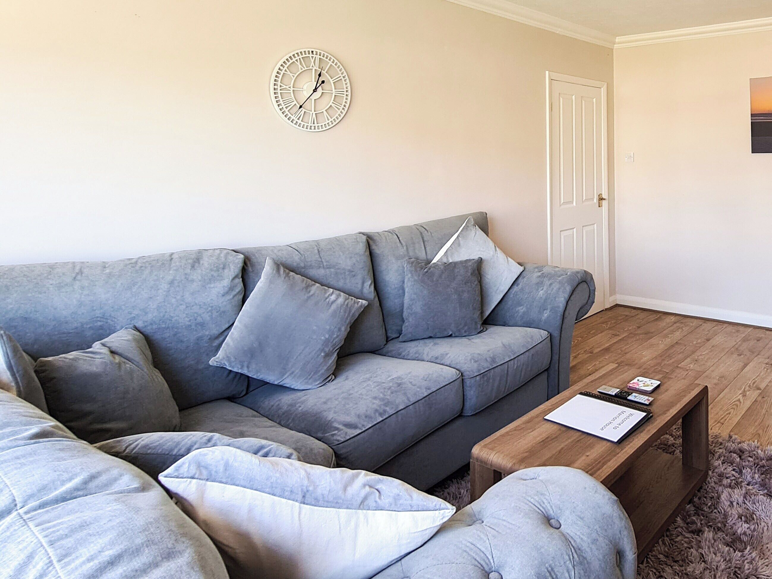 Un salón con un sofá gris, varios cojines, una mesa de centro de madera y un reloj de pared.